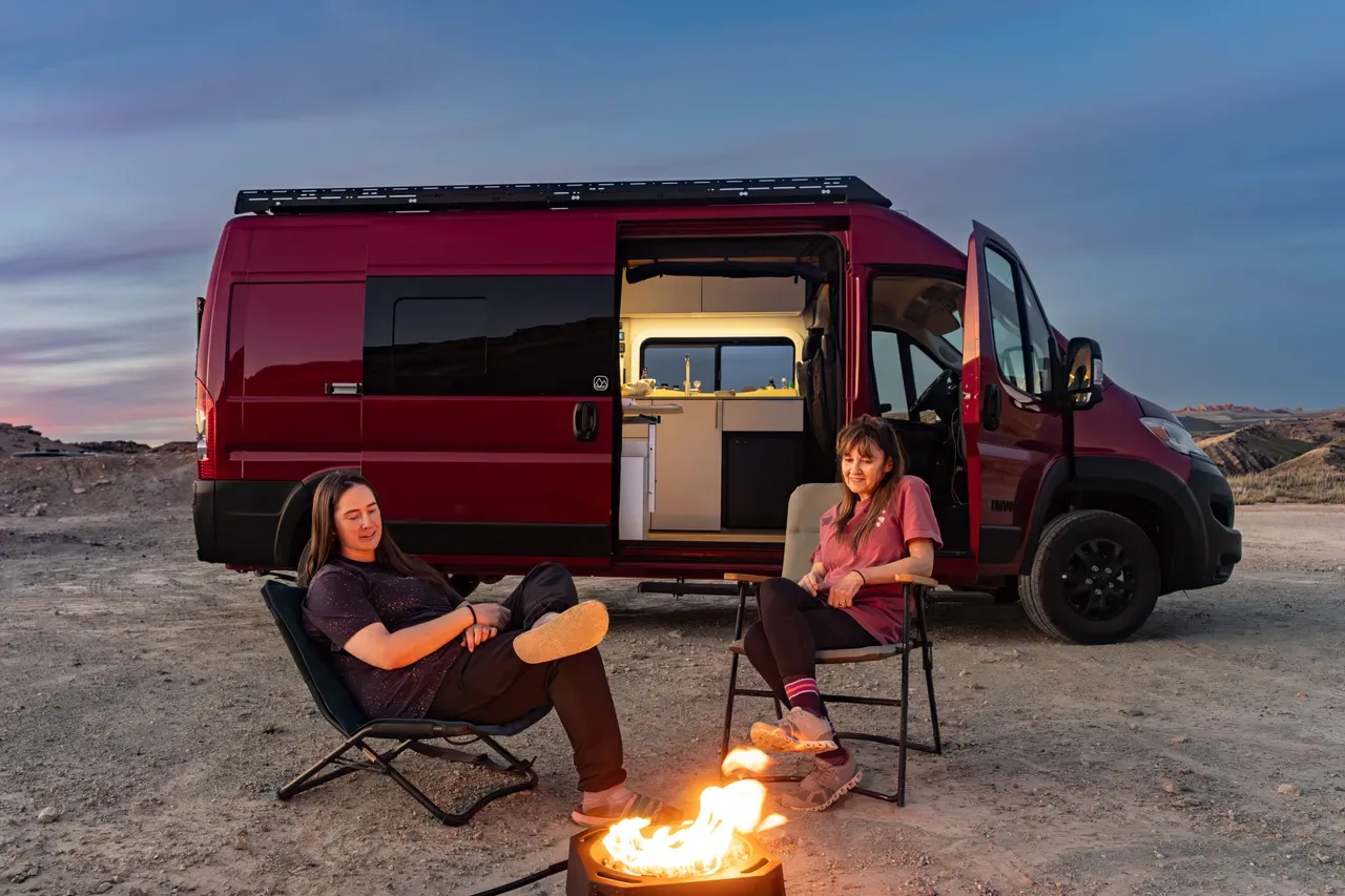 Two women sitting by a campfire outside a red LV5 camper van at dusk