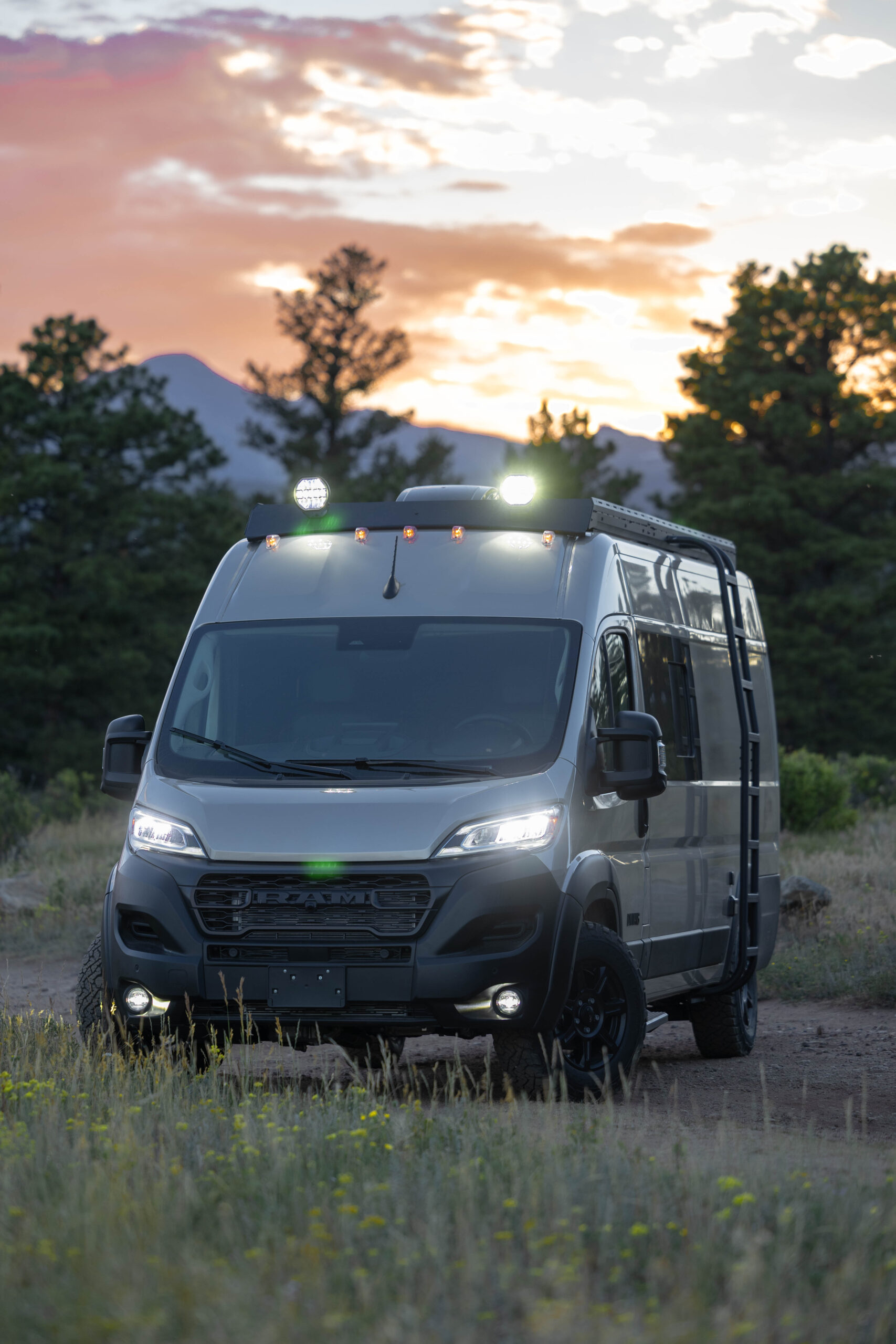 A 2027 DM Vans LV7 camper van at dusk, lights on, pines and a sunset behind